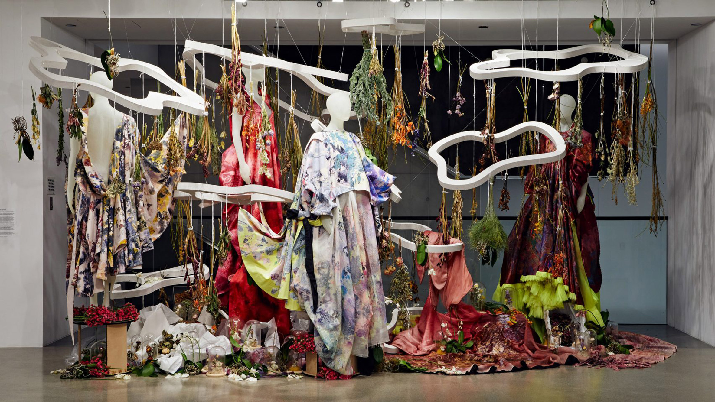 A photograph of mannequins wearing various garments in multicoloured tones. Above them are white circular installations hanging from the ceiling.