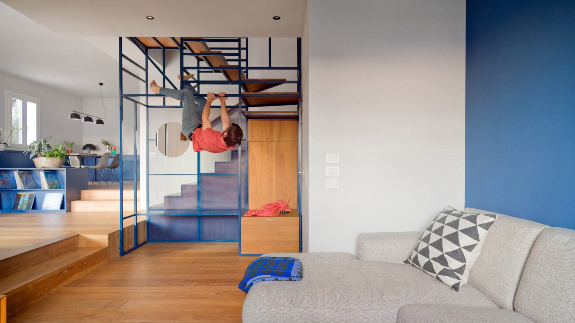 A photograph of an interior space, with a grey sofa and white and blue walls. There is a blue staircase in the centre of the image which has a square, geometric frame and a built in climbing wall, which a person can be seen interacting with in the image.