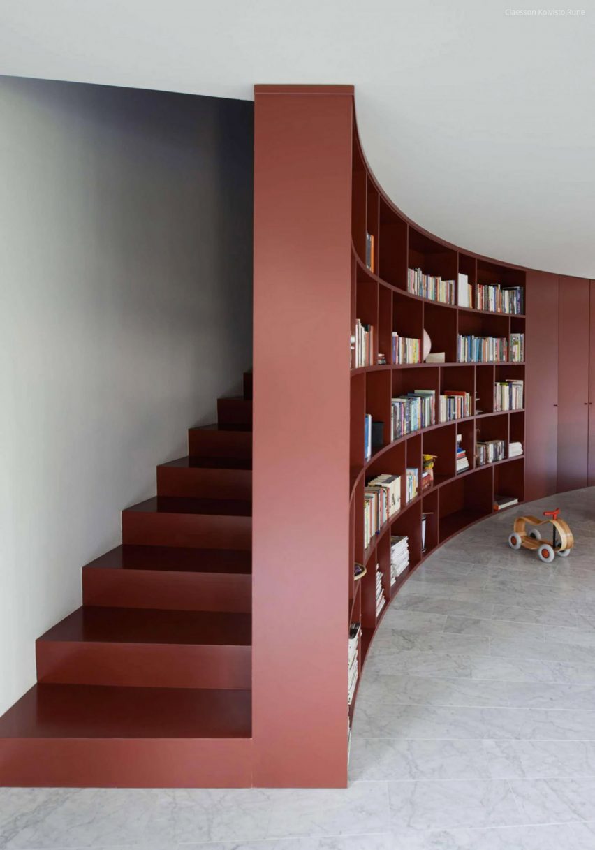 A photograph of a curved red bookshelf with a staircase behind it.