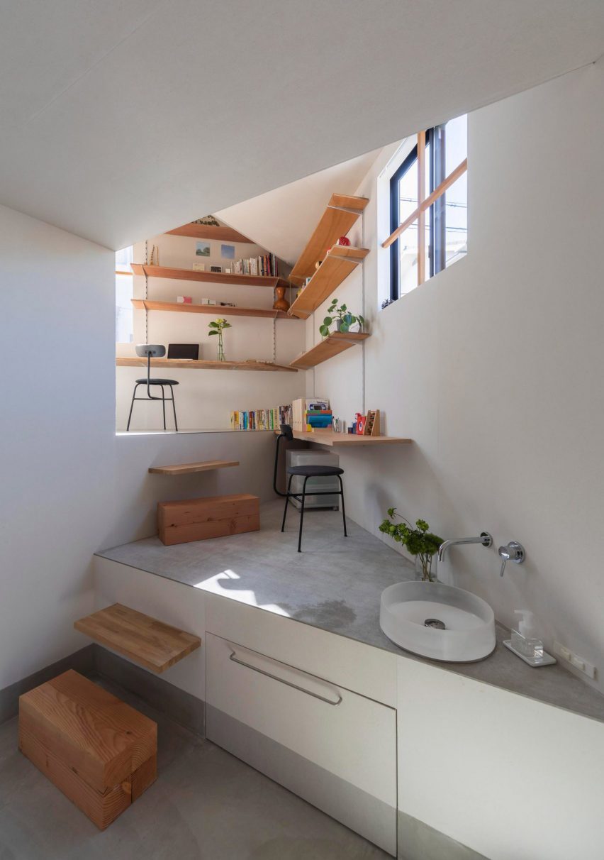 A photograph of an interior space in tones of white and grey, with brown wooden shelving throughout that double as stairs. There are also black chairs in the space and a sink.