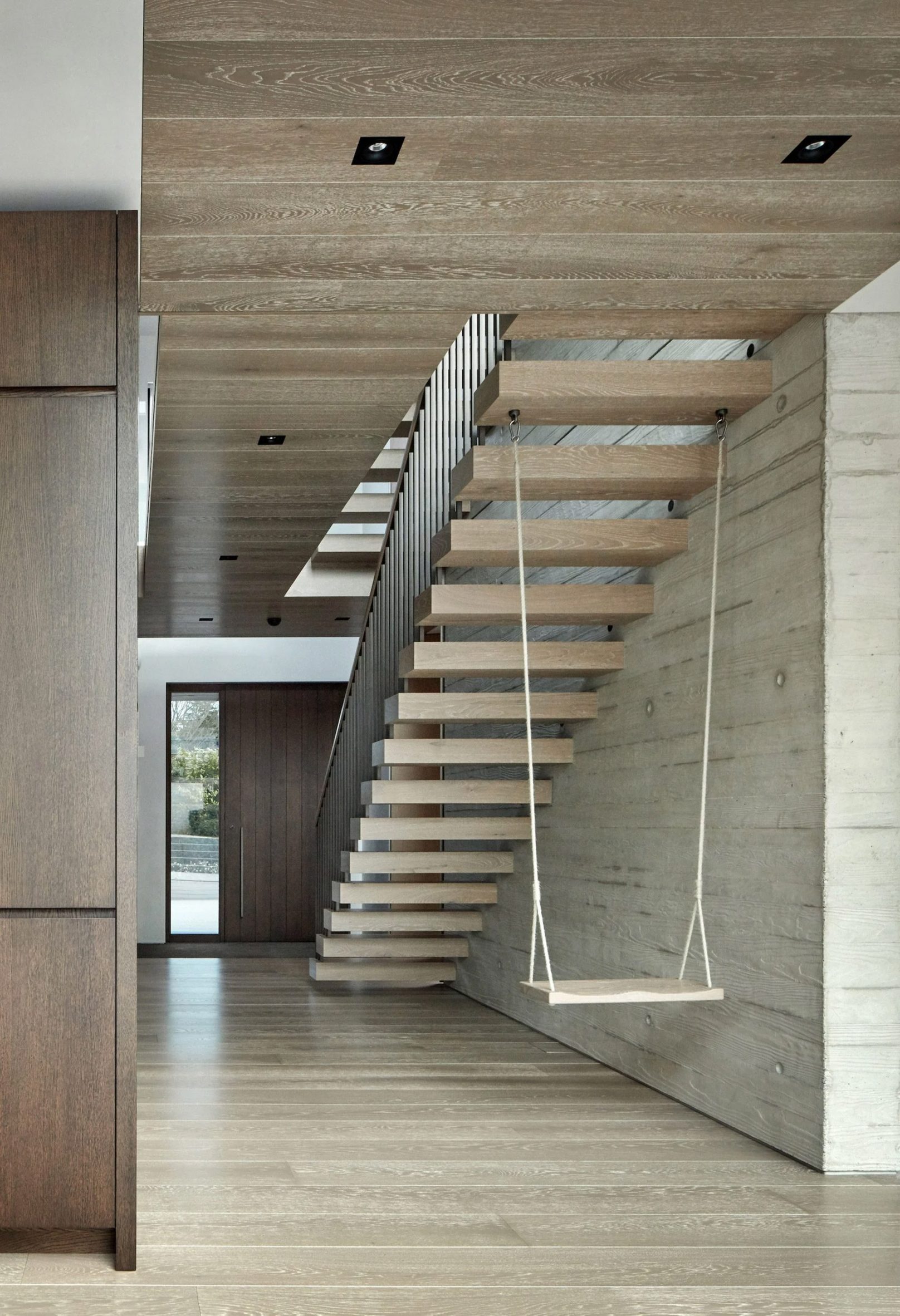 A photograph showing the underneath of a wooden floating staircase in tones of brown, with a white swing hanging from one of the stairs.