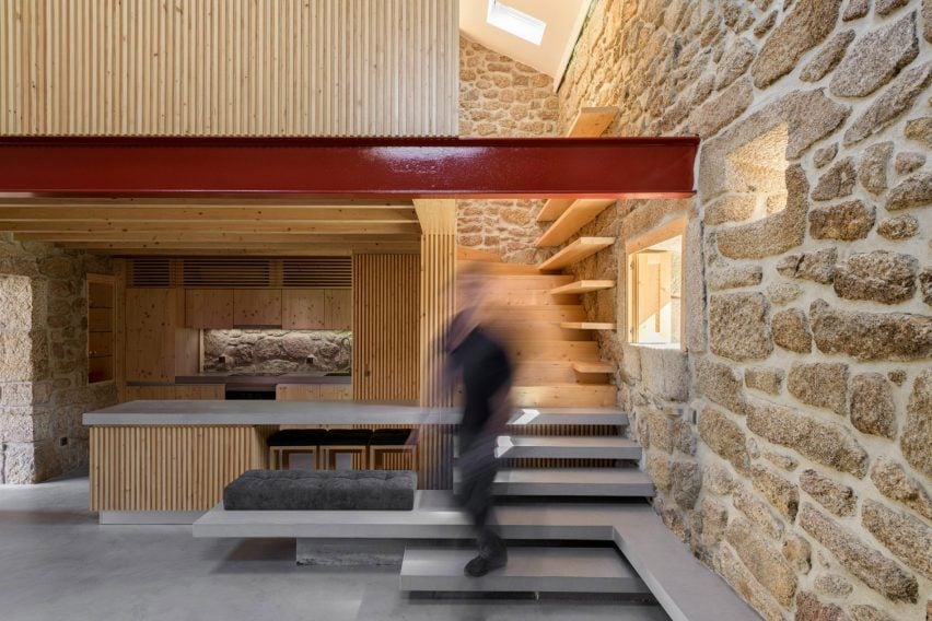 A photograph of an interior space with various stacked surfaces in tones of grey and brown. There a stone wall on the right with a red beam attached to it, and a person captured in a blur walking through the space.