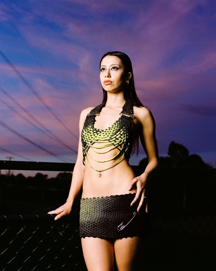 A photograph of a person wearing woven garments in tones of green and black. Behind them is a sunsetting sky in tones of pink and blue.