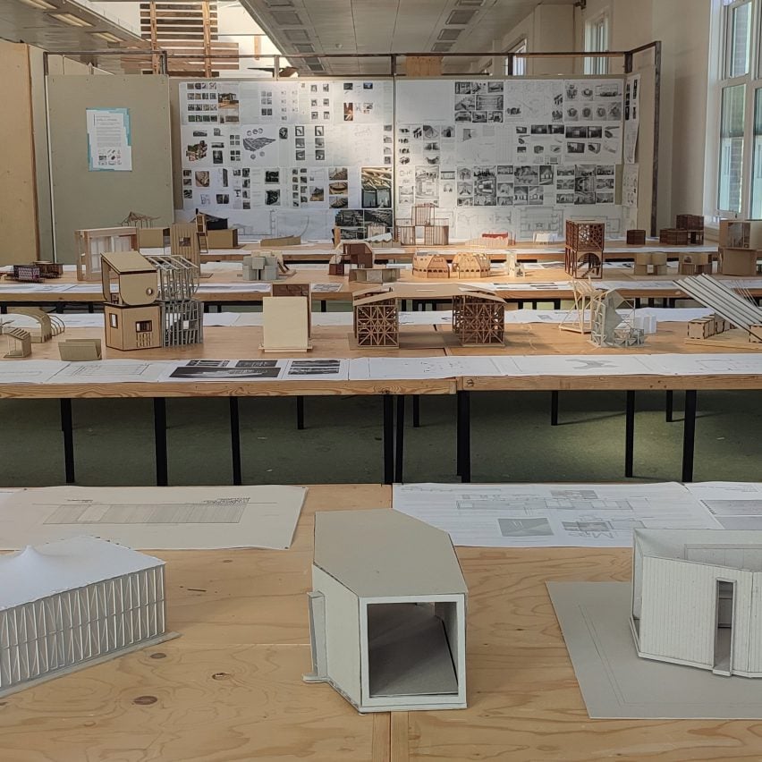 A photograph of a classroom with multiple tables in it which have different student architecture models on top of them. Towards the back of the classroom is a noticeboard with various images attached to it.