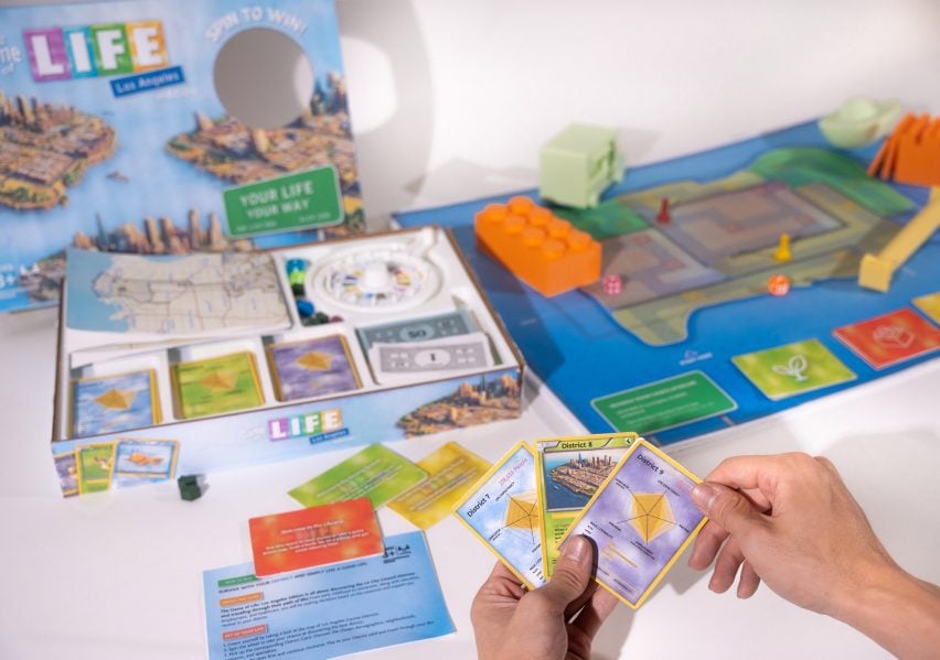 A photograph of a board game featuring tones of blue, orange, red and green. A person's hands can be seen holding three cards.