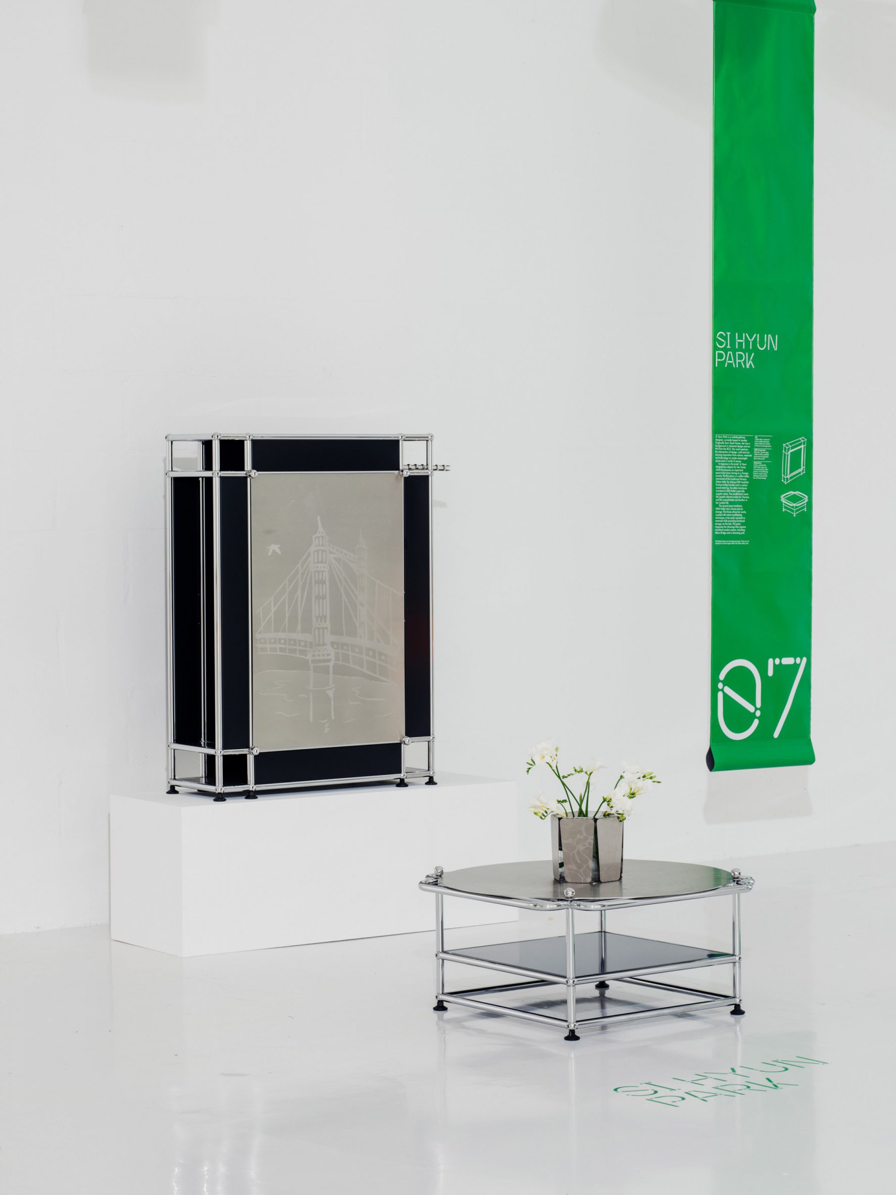 A photograph of a frame in tones of black, structured by silver beams, and a coffee table in the same colours with a vase of flowers on it. On the right there is a rectangular banner hanging in tones of green with white text on it.