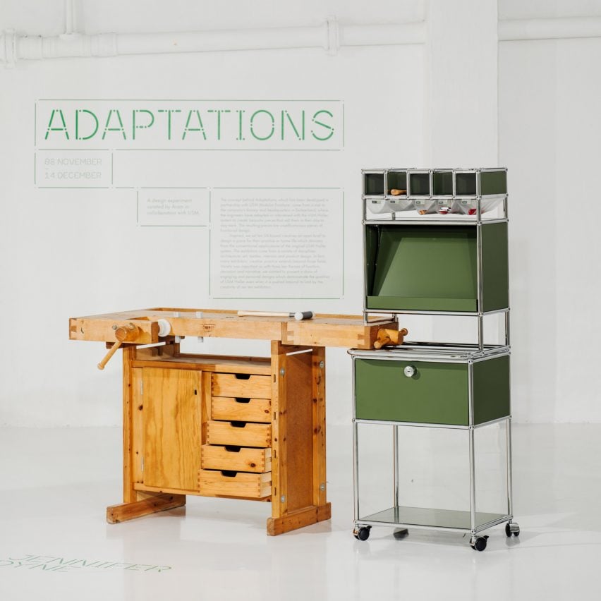 A photograph of a brown wooden workbench with a storage unit built into it. There is another storage unit beside it in tones of green, structured by silver beams. Behind them both is a white wall with green text on it.