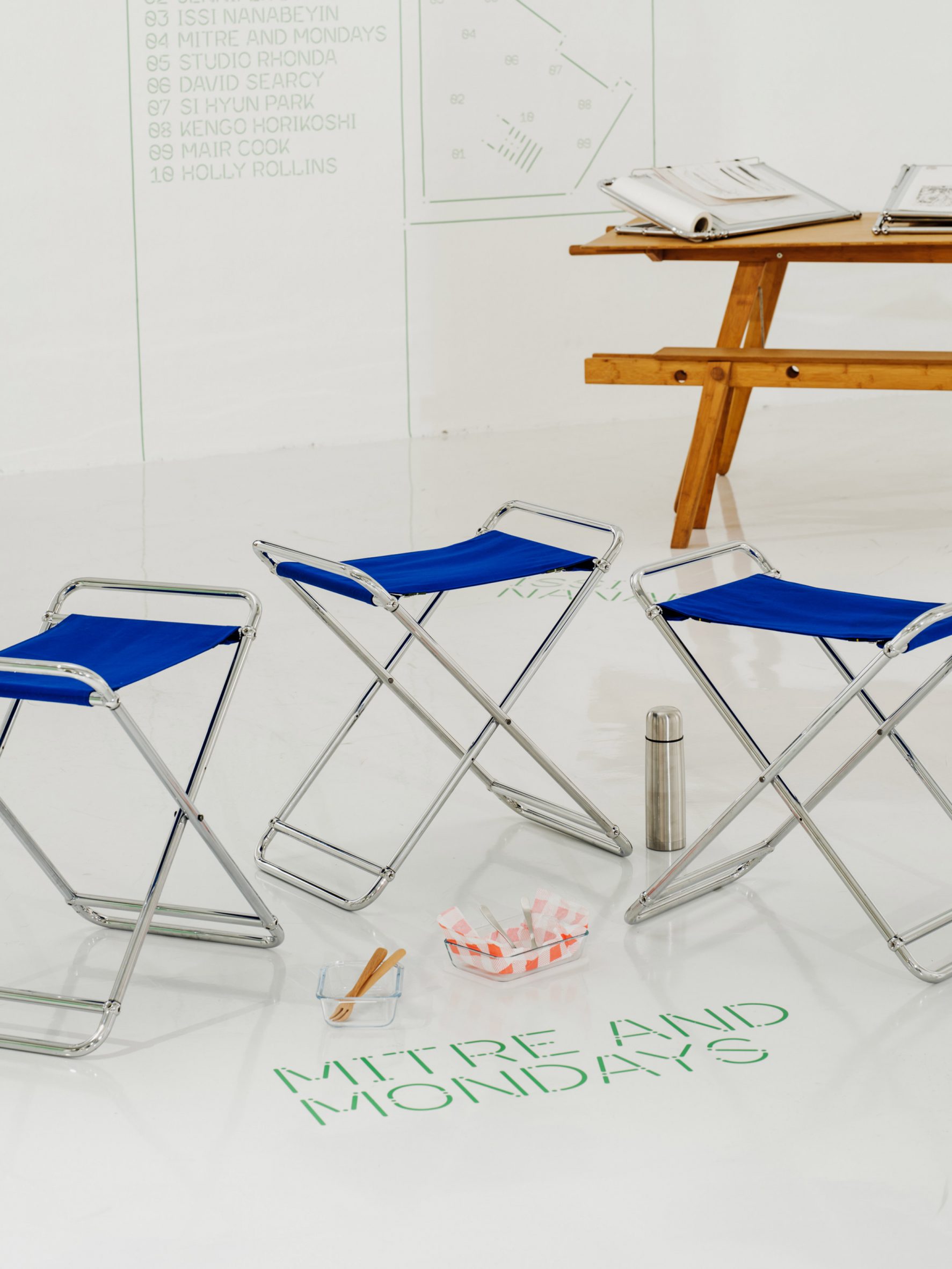 A photograph of three blue stools with structured by silver beams, with a brown wooden picnic table behind them.