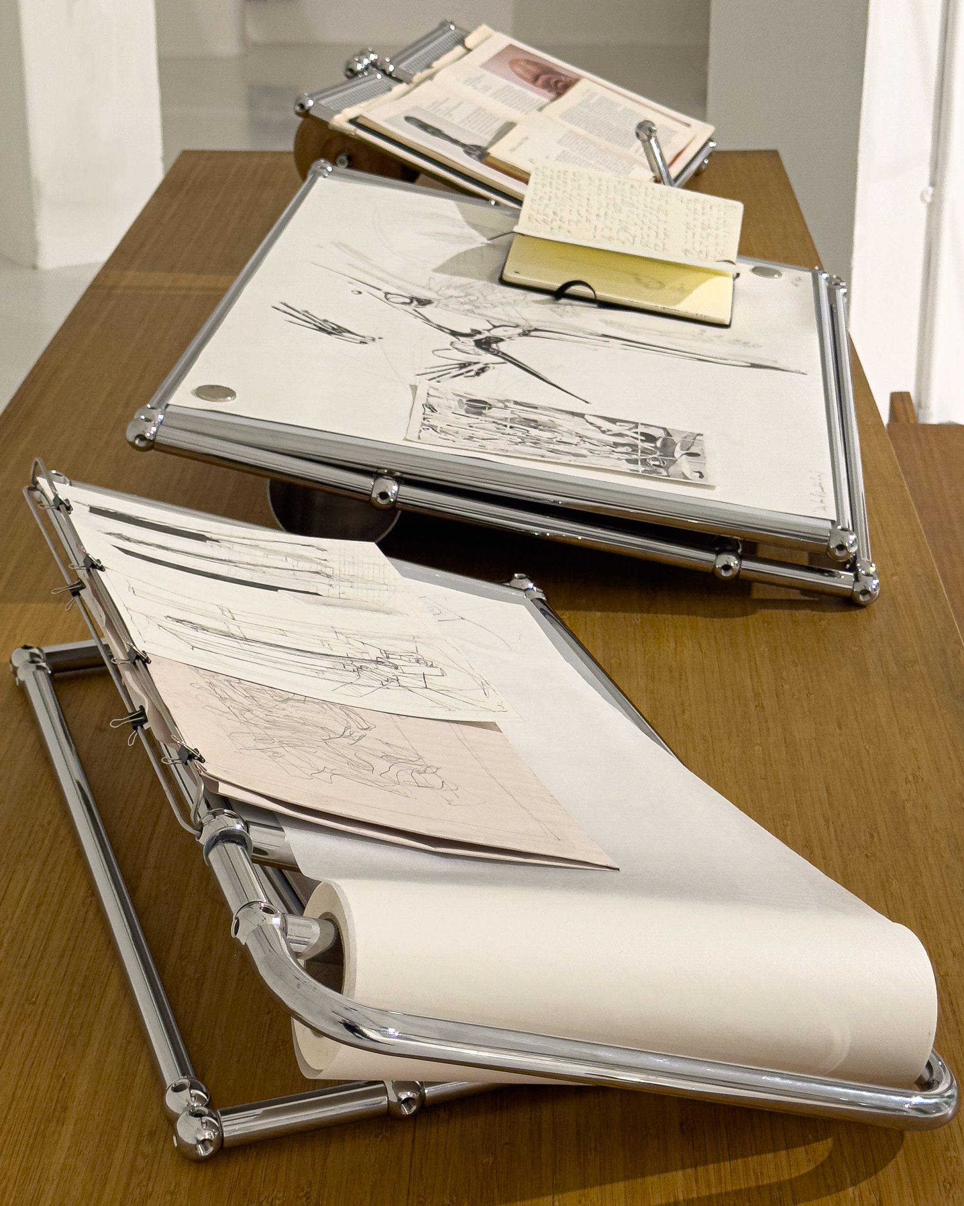 A photograph of silver drawing boards with white paper on them, displaying pencil drawings.