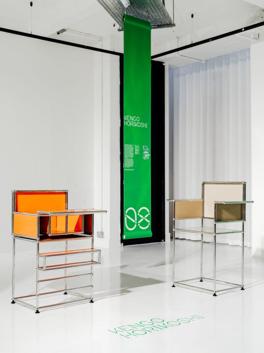 A photograph of two chair-like objects, structured by silver beams. One is orange and one beige. Between them is a hanging rectangular green banner with white text on it.