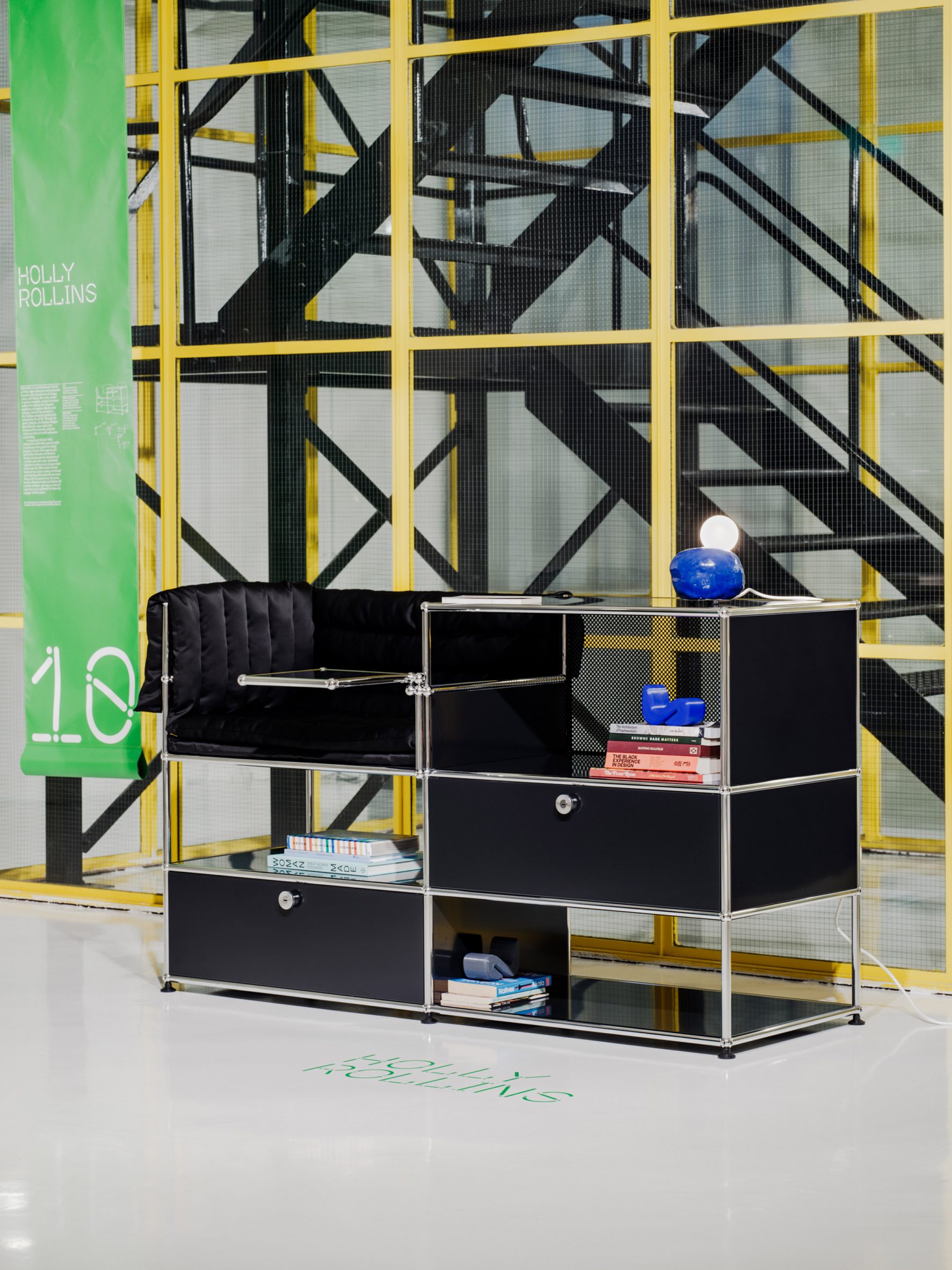 A photograph of a storage unit in tones of black and silver, against a glass wall with yellow beams.