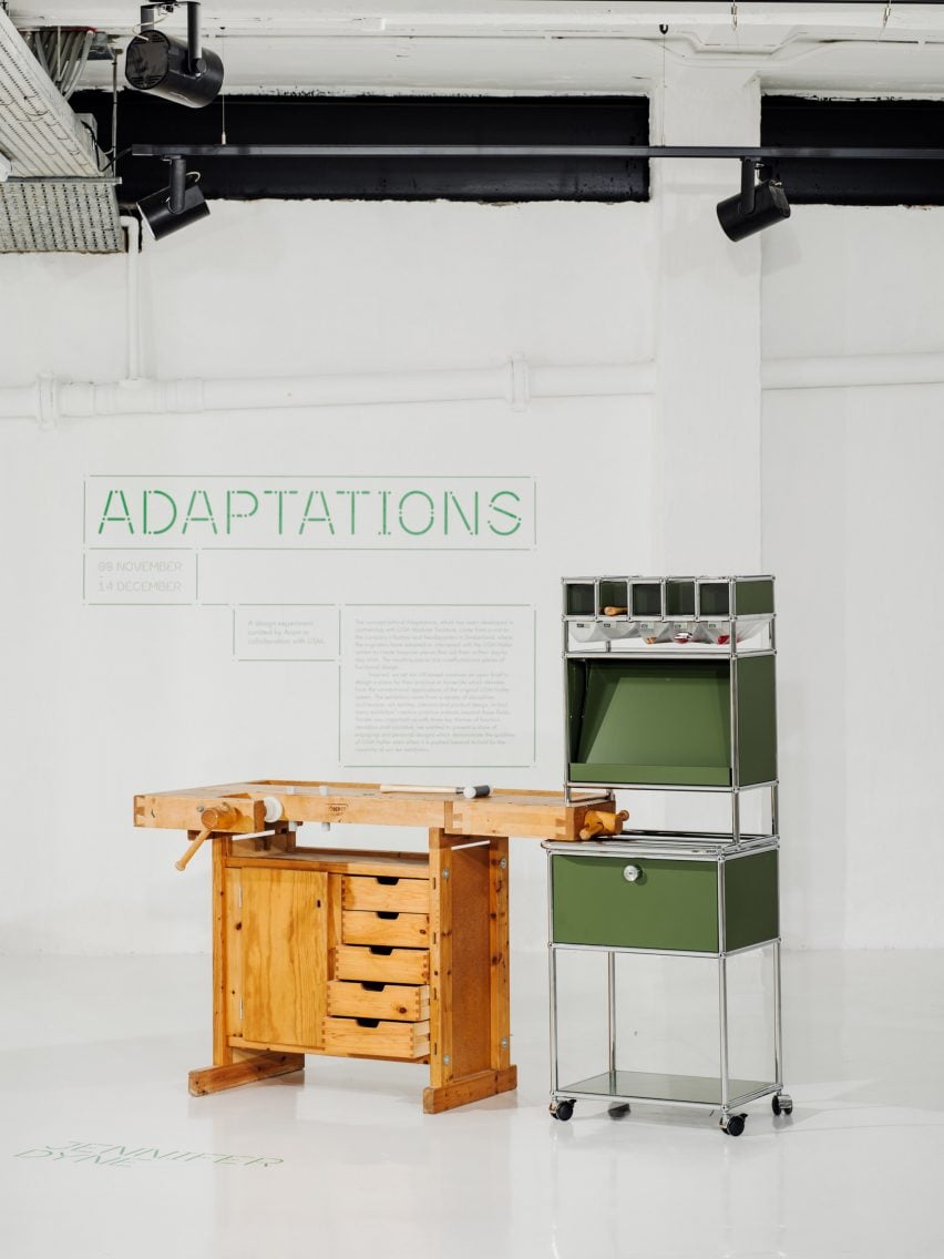 A photograph of a brown wooden workbench with a storage unit built into it. There is another storage unit beside it in tones of green, structured by silver beams. Behind them both is a white wall with green text on it.