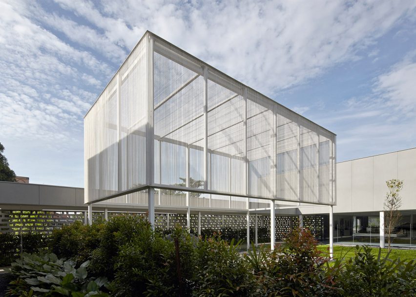 A photograph of a building comprised of white beams, held up by legs, among greenery. There is a blue sky above.