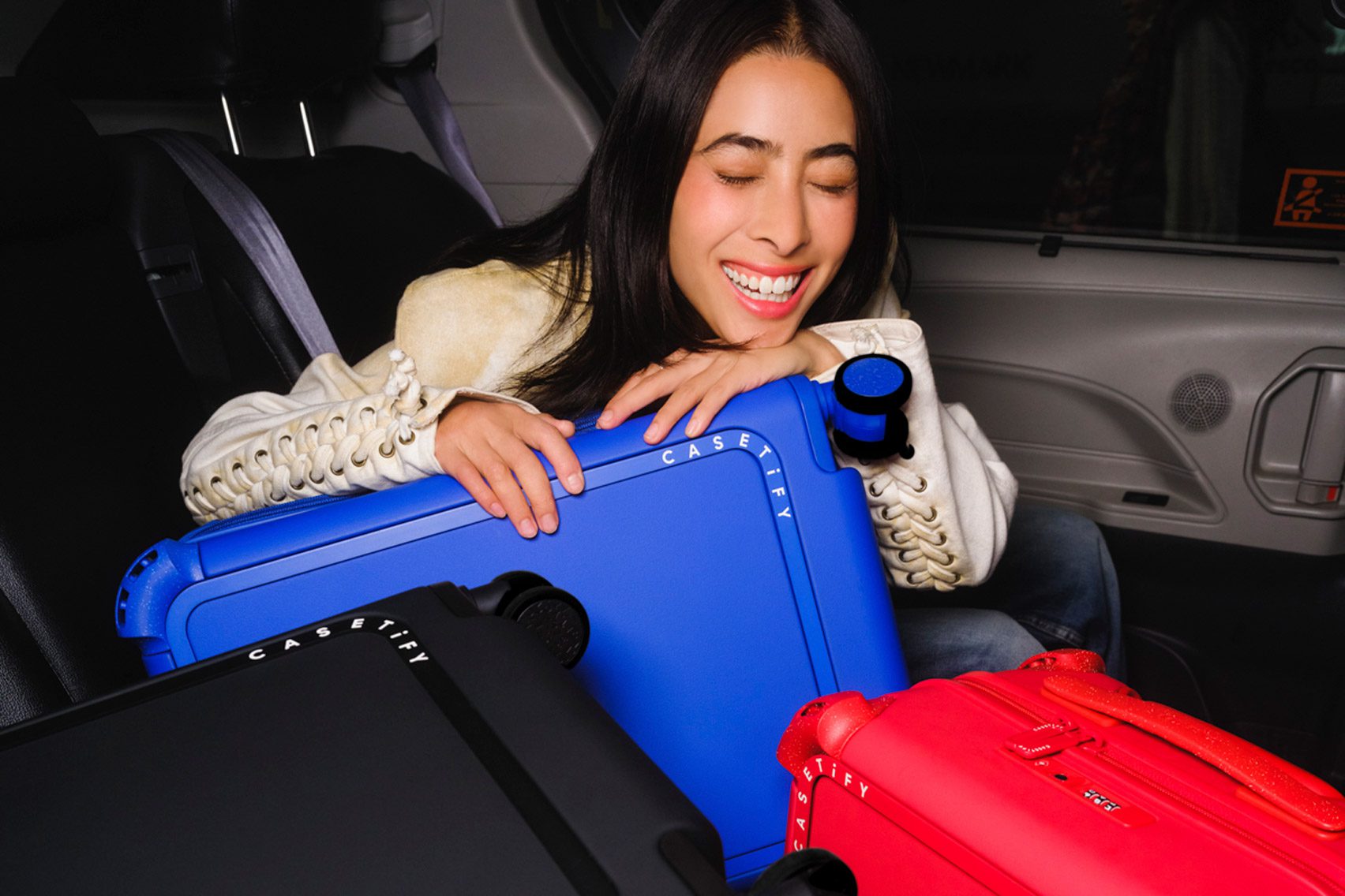 Photo of a woman looking happy as she rests on three brightly coloured Casetify suitcases in the back of a car