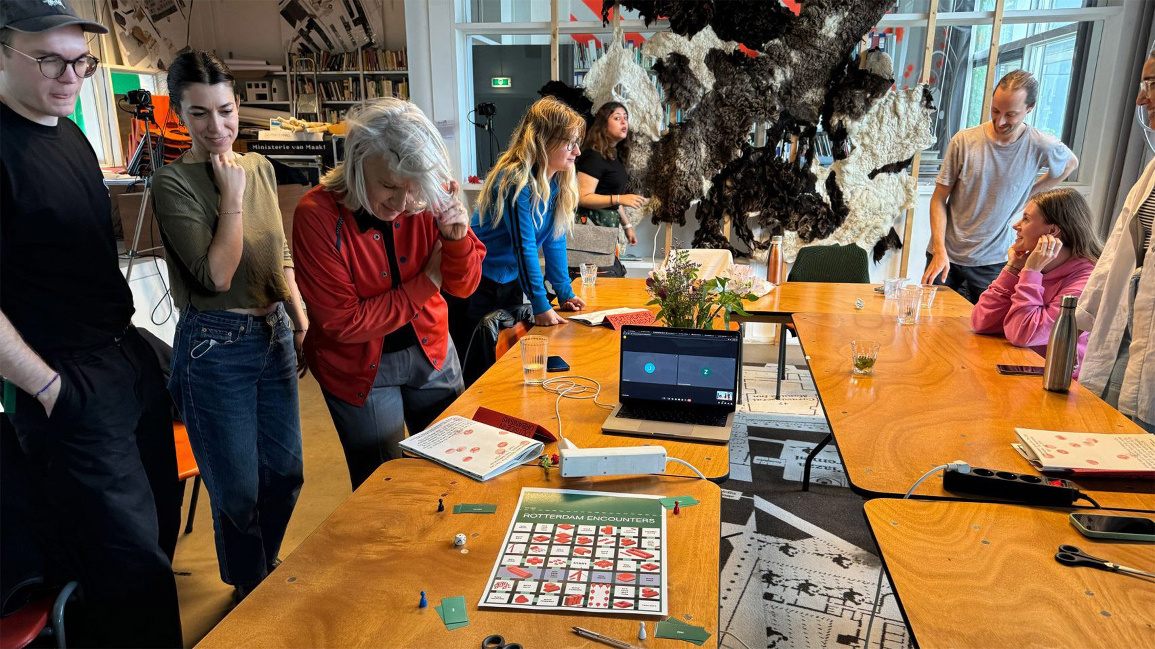 A photograph of students and tutors discussing their work, which is laid out on a brown wooden table.