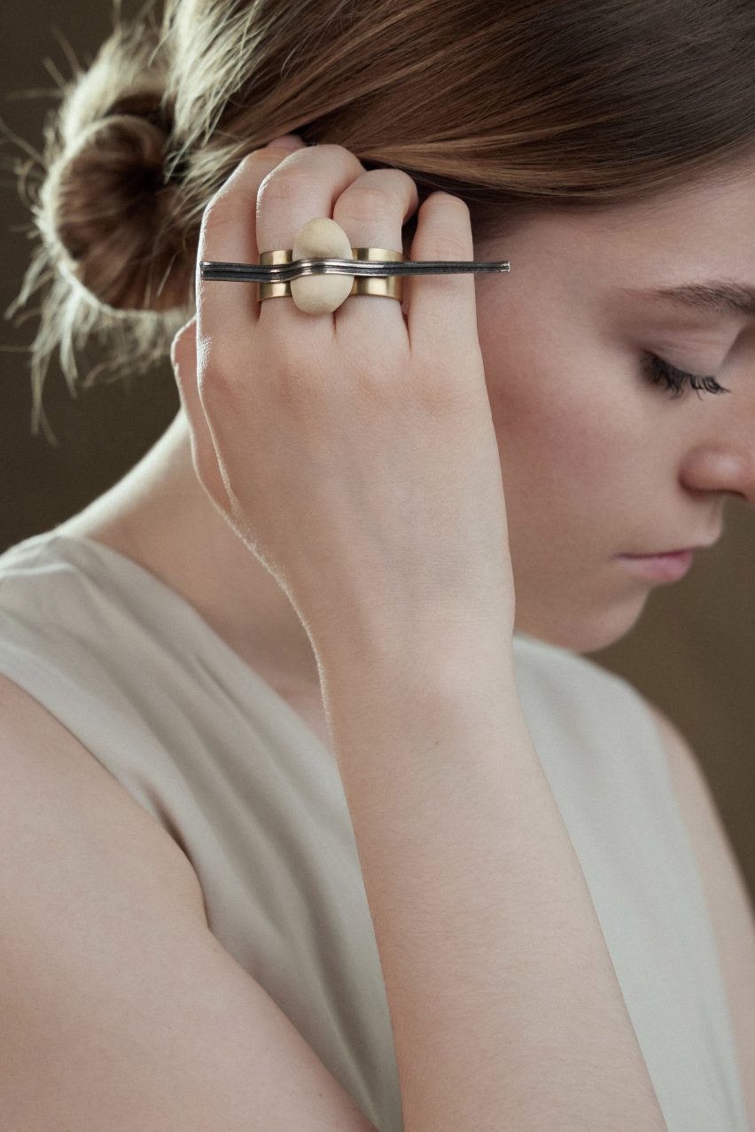 Close-up photo of a woman's head as she tucks hair behind her ear. She is wearing a chunky ring by Cindy Xinyi Wu across two fingers that features a ball of baked dough flattened in the middle by a long, textured metal element that spans the width of the hand