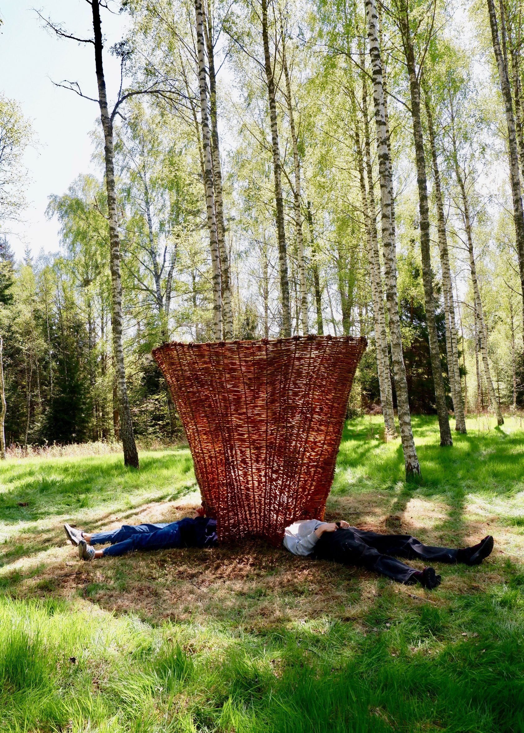 A photograph of a brown woven structure within a green forest. There are two people lying down next to it.