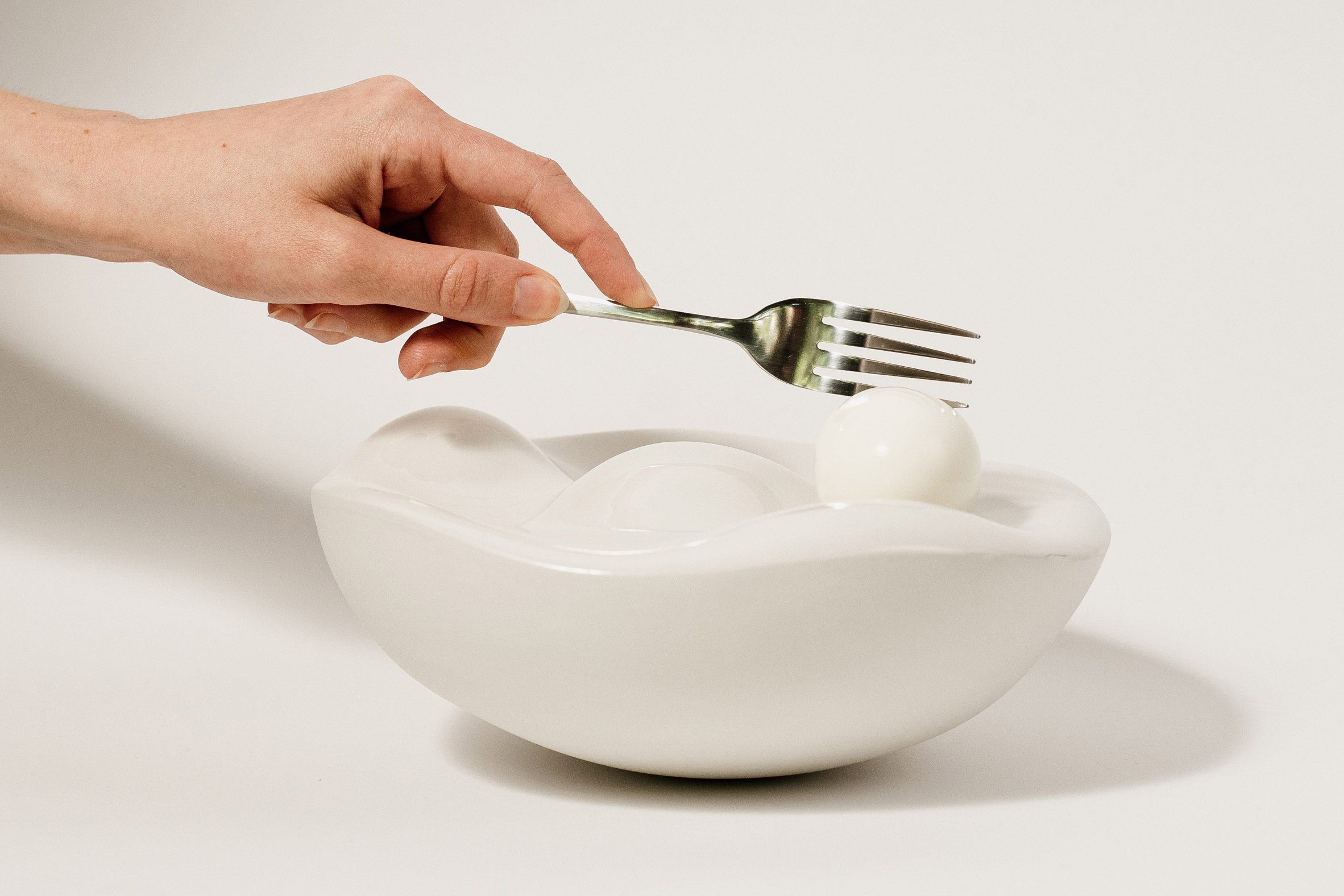 A photograph of a person's hand using a fork to eat a boiled egg, of which there are three within a white dish.