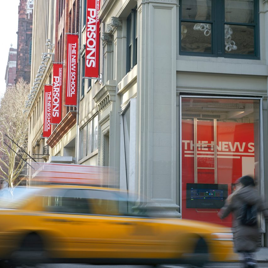 A photograph of a building in tones of grey, with red flags on its exterior. There is a moving yellow car driving in front.