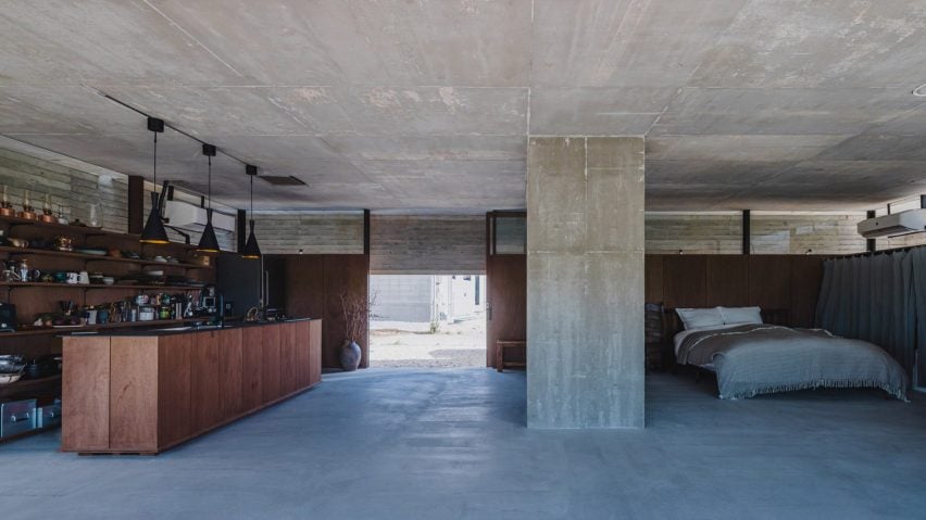 A photograph of large open-plan apartment with a brown wooden bar and bed in it. The ceiling, walls and floor are all concrete, in tones of blue and grey.