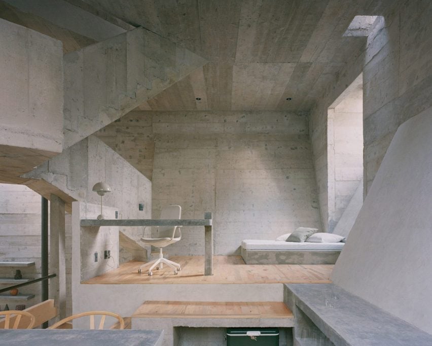 A photograph of a room that is comprised of various geometric structures, made up pf concrete and in tones of grey. There is a white chair and bed in the room.