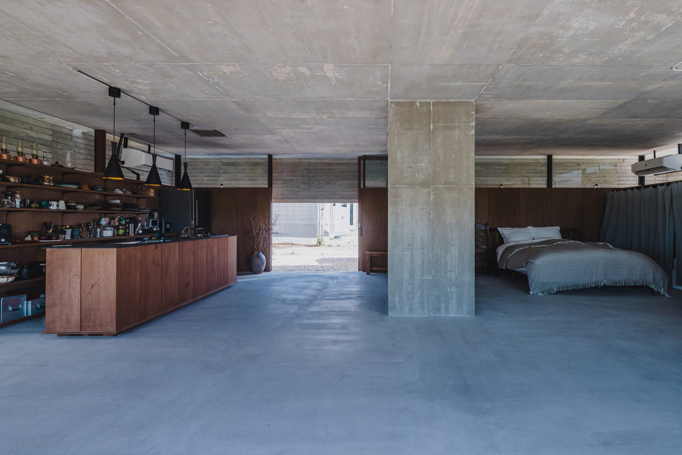 A photograph of large open-plan apartment with a brown wooden bar and bed in it. The ceiling, walls and floor are all concrete, in tones of blue and grey.