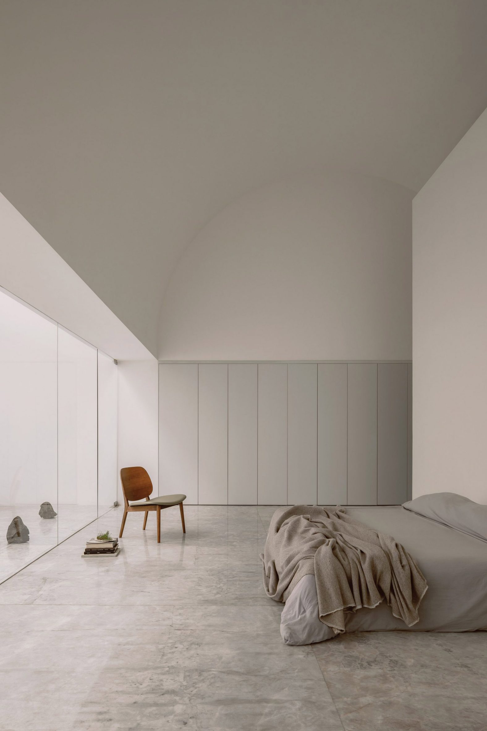 A photograph of a large room in tones of white and grey, with a bed and brown wooden chair in it. One of the walls is glass.