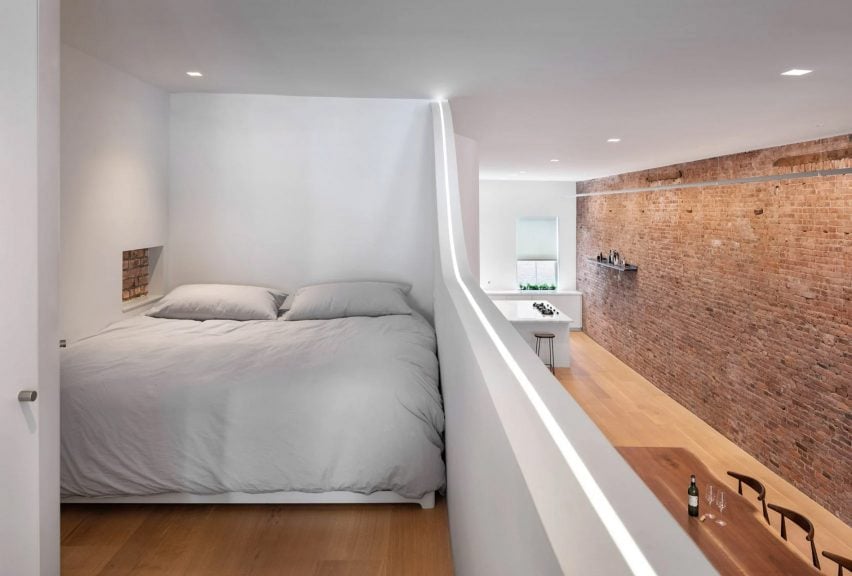 A photograph of a bed atop a white mezzanine, overlooking a brick wall and wooden floor.