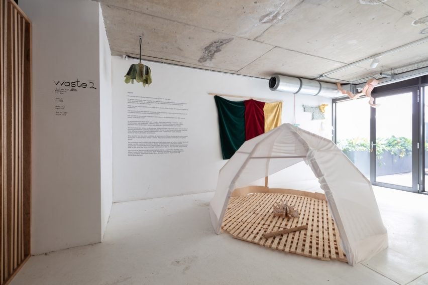 Photo of a white tent-like structure with a slatted timber base within a white exhibition space, surrounded by other objects