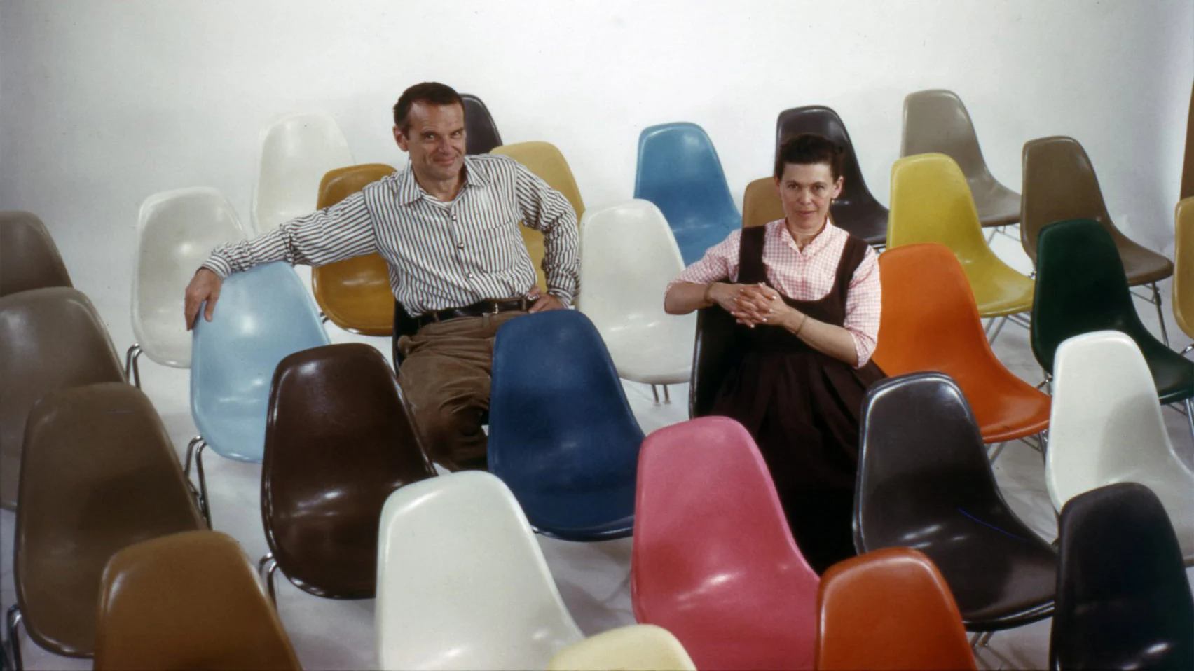 Ray and Charles Eames seated amongst group of Shell chairs 