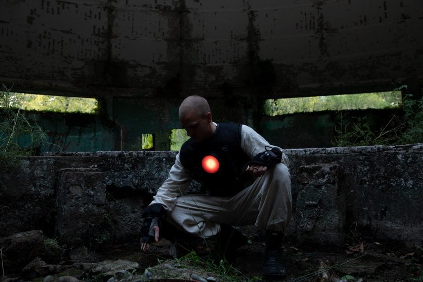 Photo of a man in loose beige clothing and a black vest with a glowing red disc over the heart crouching down to touch the earth in the middle of a ruined building overrun with plants