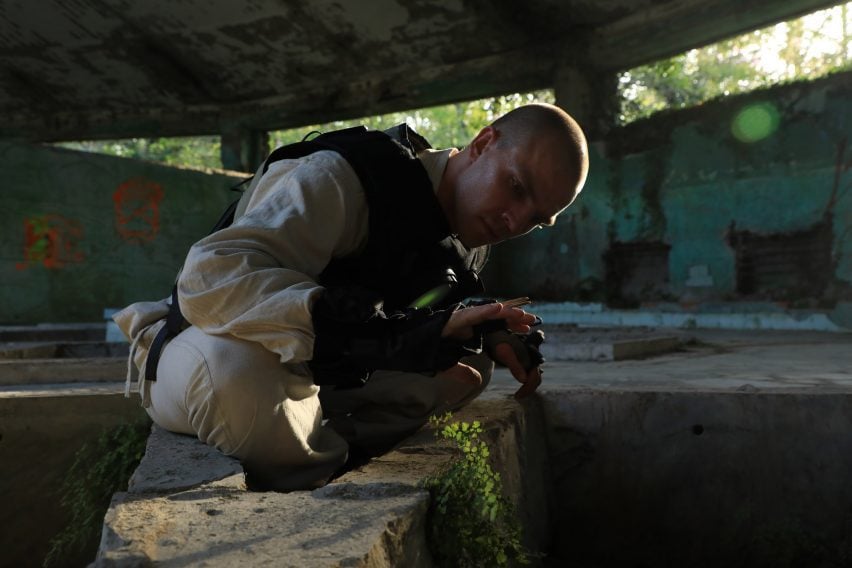 Photo of a man exploring a ruined old building that has been taken over by plants while wearing a glove and vest packed with sensors. His hand hovers over a plant as he looks at it intently