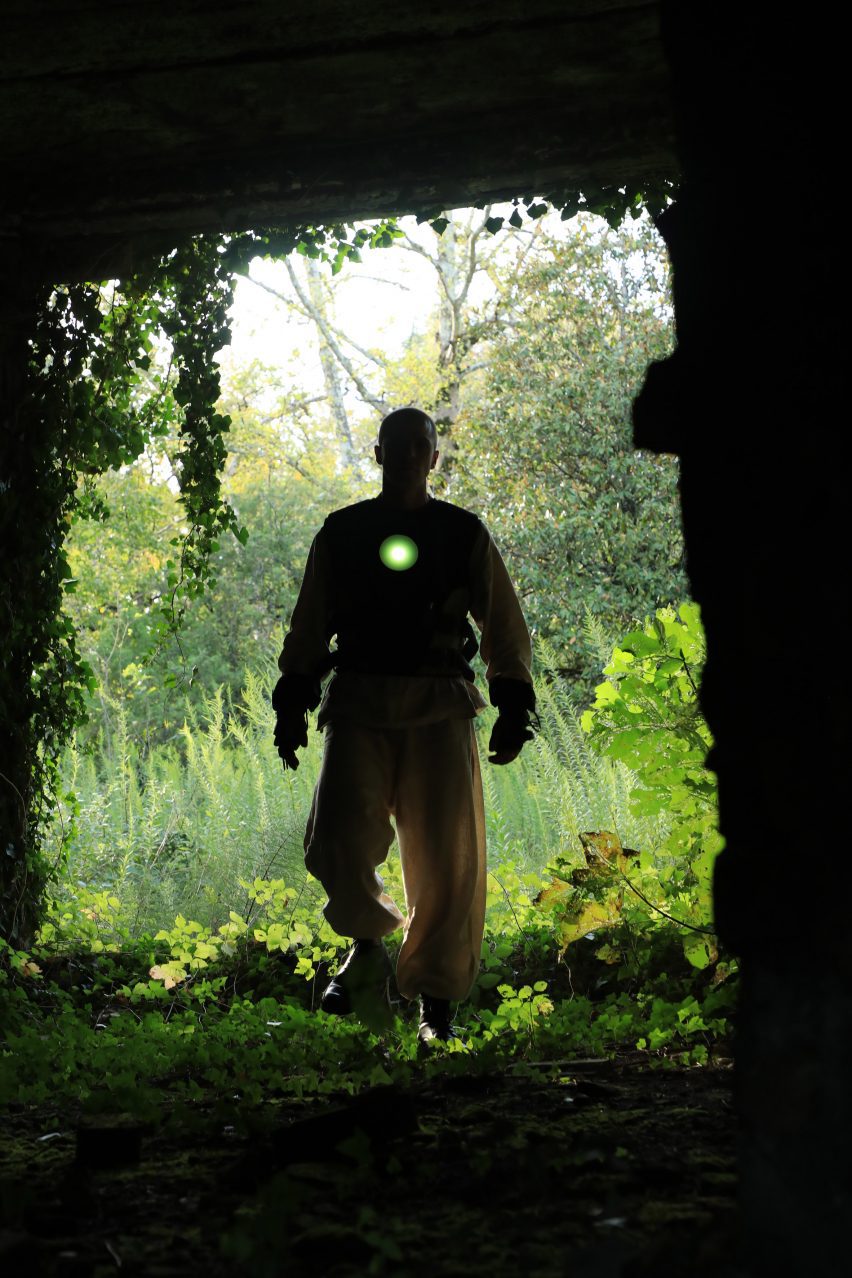 Photo of a man silhouetted through an entrance to a structure in a lush, green forest. He wears an outfit with a glowing green disc over the heart