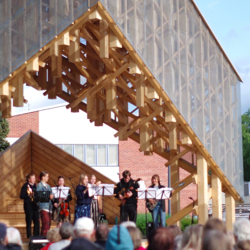 A photograph of a band playing within a wooden structure outdoors. There are people watching.