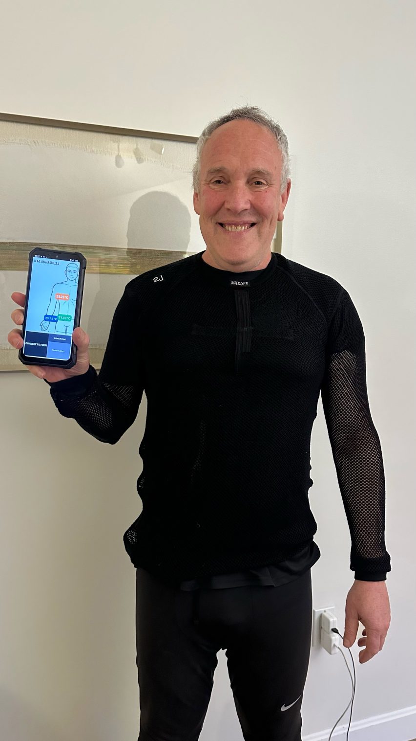 Photo of Professor Yoel Fink wearing a simple black shirt and leggings woven with the fibre computer and holding a smartphone showing data mapped onto a figure of the human body