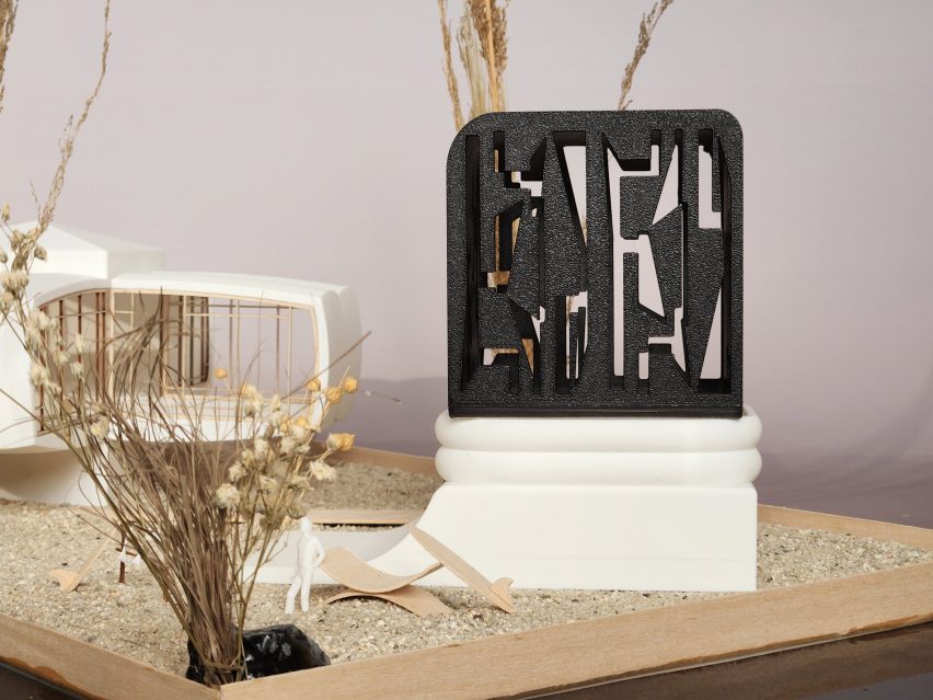 A photograph of an architectural model, in tones of white and black, atop a beige sanded surface.