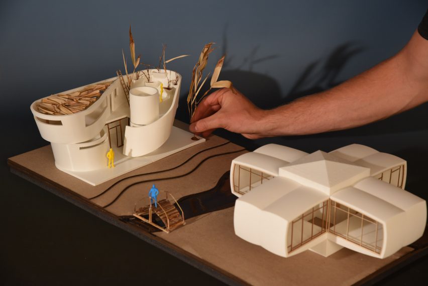 A photograph of a person's hand interacting with an architectural model in tones of white, atop a beige surface.