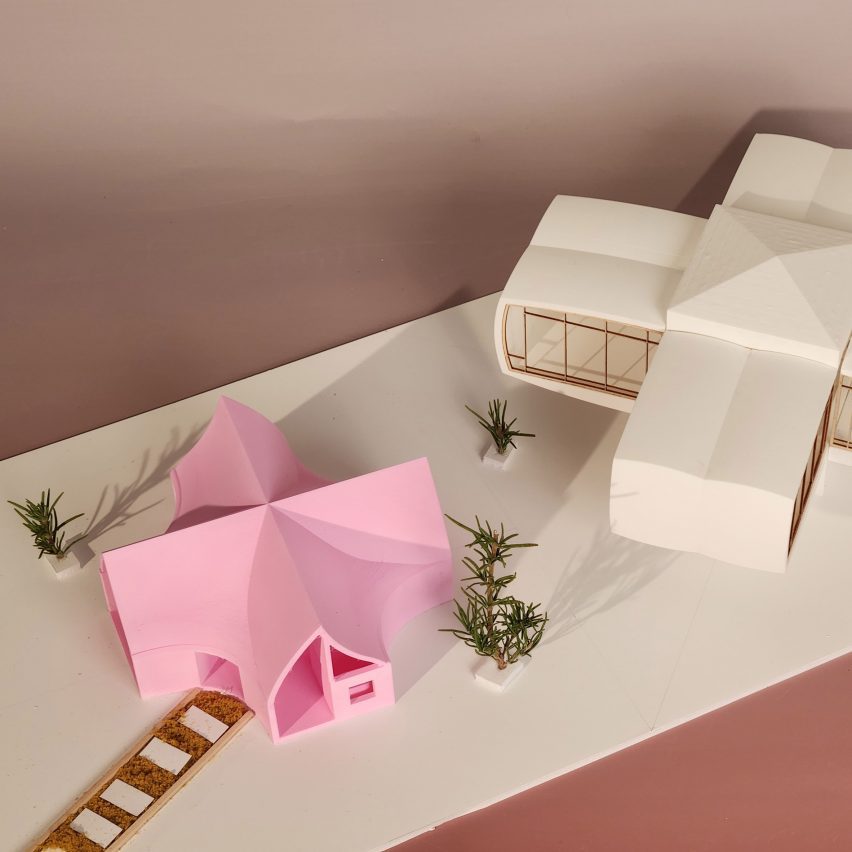 A photograph of an architectural model in tones of white and pink, atop a white surface. There is a brown backdrop.