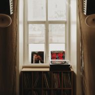 Photograph of a window framed by linen curtains with a vinyl cabinet and a turntable. On either side of the window hands two bespoke speakers.