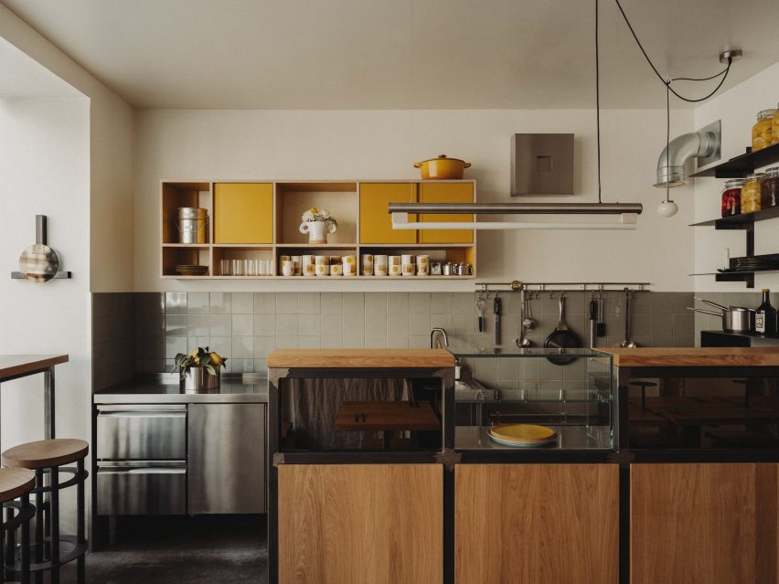Photograph of an open-plan kitchen and bar area located at the front of the restaurant interior. The area is made up of an oakwood bar, steel kitchen basins and yellow plywood shelves.