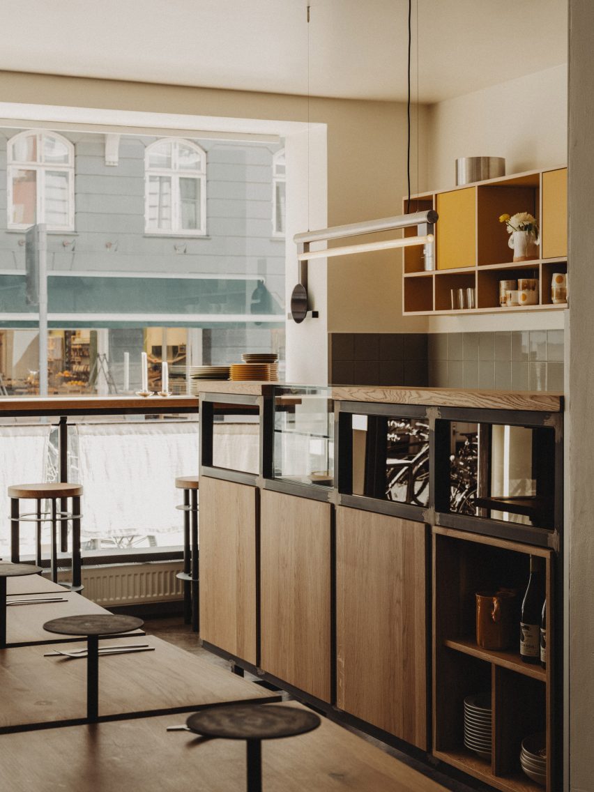 Photograph showing a minimalist restaurant interior with an open-plan design and natural oak furnishings. A bar area is located at the front of the restaurant, which overlooks the street through a large glass facade.