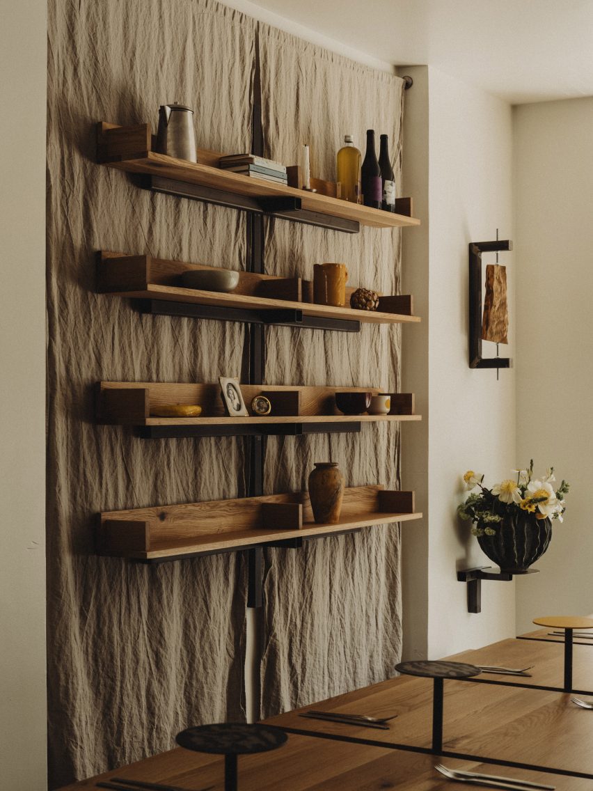 A photograph showing earthy, natural linen hanging from the walls, with floating oakwood shelves displaying miscellaneous items such as vases, books and bottles.