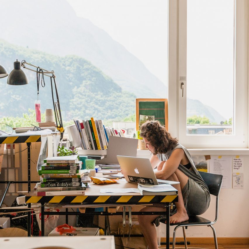A photograph of a student sat working at a desk beside a large window.