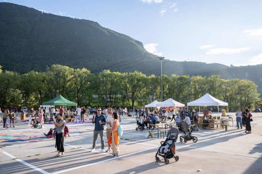 A photograph of a grey concrete ground in front of a mountain and green trees. There are multiple people in the space.