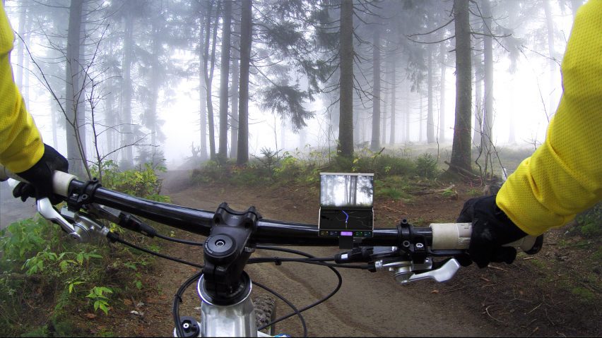 Photo of a bike cycling on a forest trail, viewed from the POV of the rider, with a mini dual-screen device attached to the handlebars