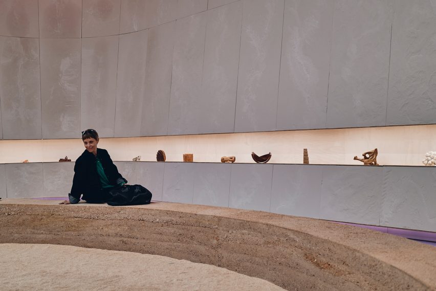 Photo of an exhibition space showing a woman sitting on a rammed earth bench seat that curves around, with sand in front and a display of objects along one linear shelf behind her
