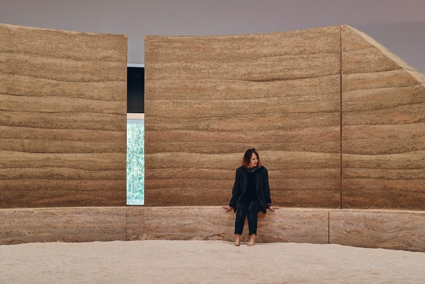 Photo of a woman sitting on a rammed earth bench in front of a semi-circular of the same material with her bare feet in sand within the Australian pavilion