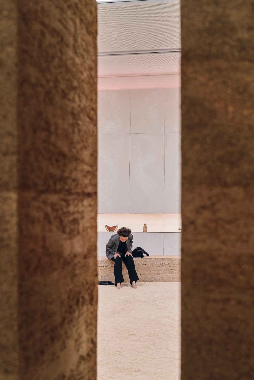 Photo looking through a clean-lined gap in a rammed-earth strcuture at a woman as she sits on bench seating and digs her bare feet into sand