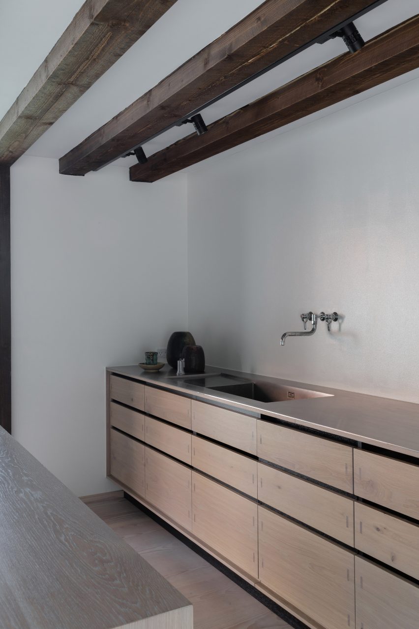 Wooden kitchen units beneath wall-mounted metal tap in a room with wooden ceiling beams
