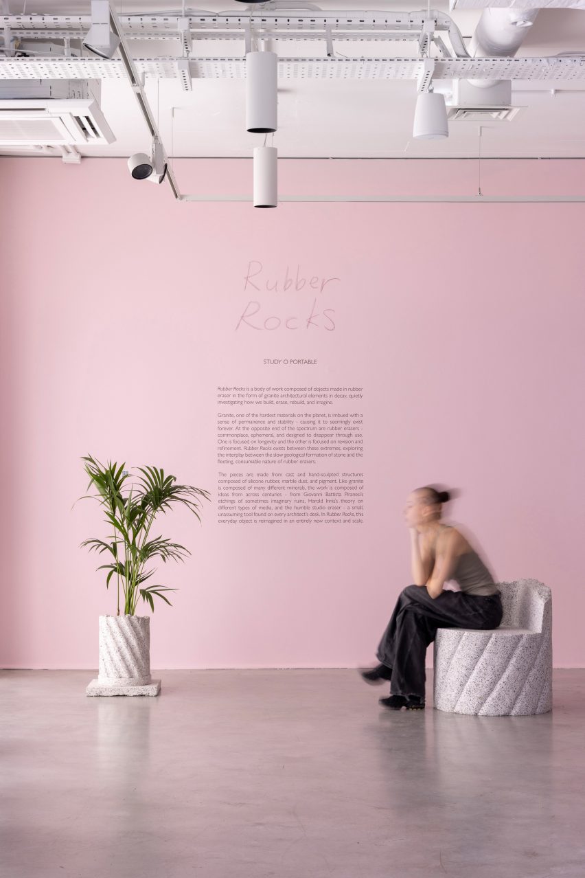 Photograph of the exhibition's explanation, which has been printed on a light pink wall, with a planter from the furniture collection and a woman sat on a stool from the collection.